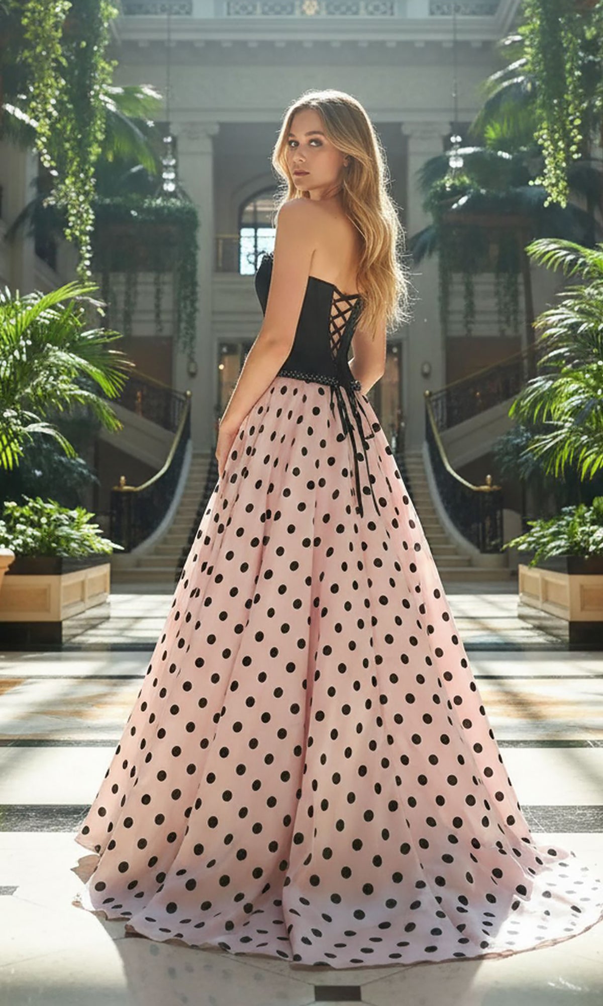 A woman in the Amelia Couture 8071 long prom dress stands in an elegant indoor garden, looking over her shoulder. The strapless black and pink polka dot a-line gown creates a striking, graceful silhouette.